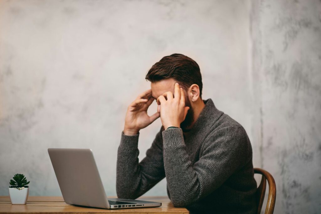 A man with dark hair is sitting at a desk in front of his laptop with his head in his hands.